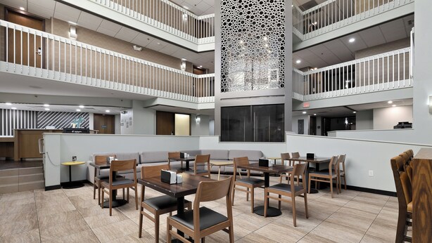 Un hall d’hôtel à plusieurs niveaux, doté d’un atrium central, de plusieurs espaces de restauration avec des tables et des chaises en bois, et d’un panneau décoratif vertical.