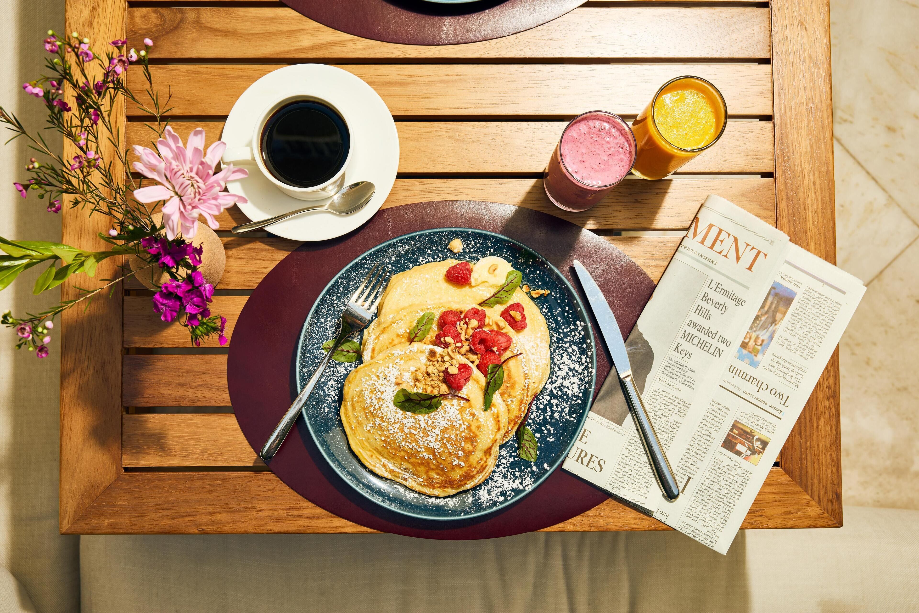 Une table en bois dressée pour le petit-déjeuner, avec des crêpes, du café, du jus de fruits et un journal.