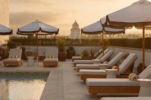 A rooftop pool area with lounge chairs and umbrellas, a large building in the background, and potted plants. - L'Ermitage Beverly Hills (Beverly Hills)