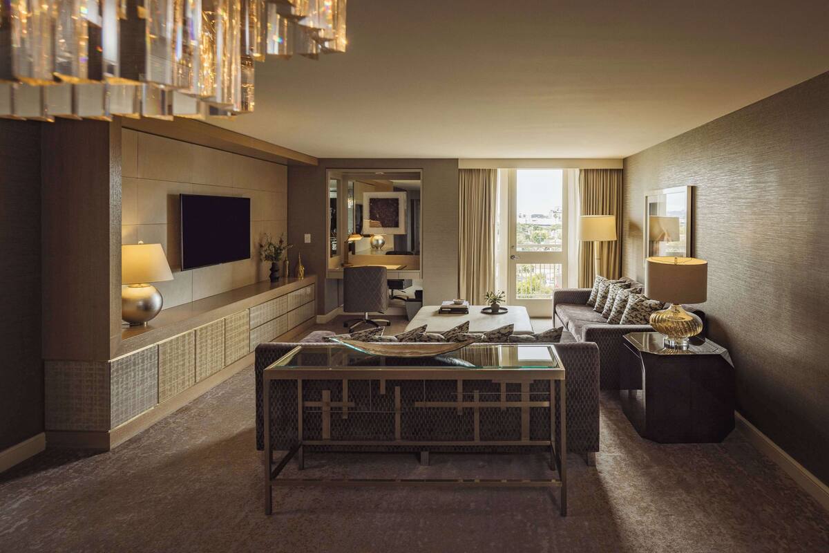 A modern hotel room with a flat-screen TV, a dining area with a glass table, a sofa, and a small side table with a lamp.