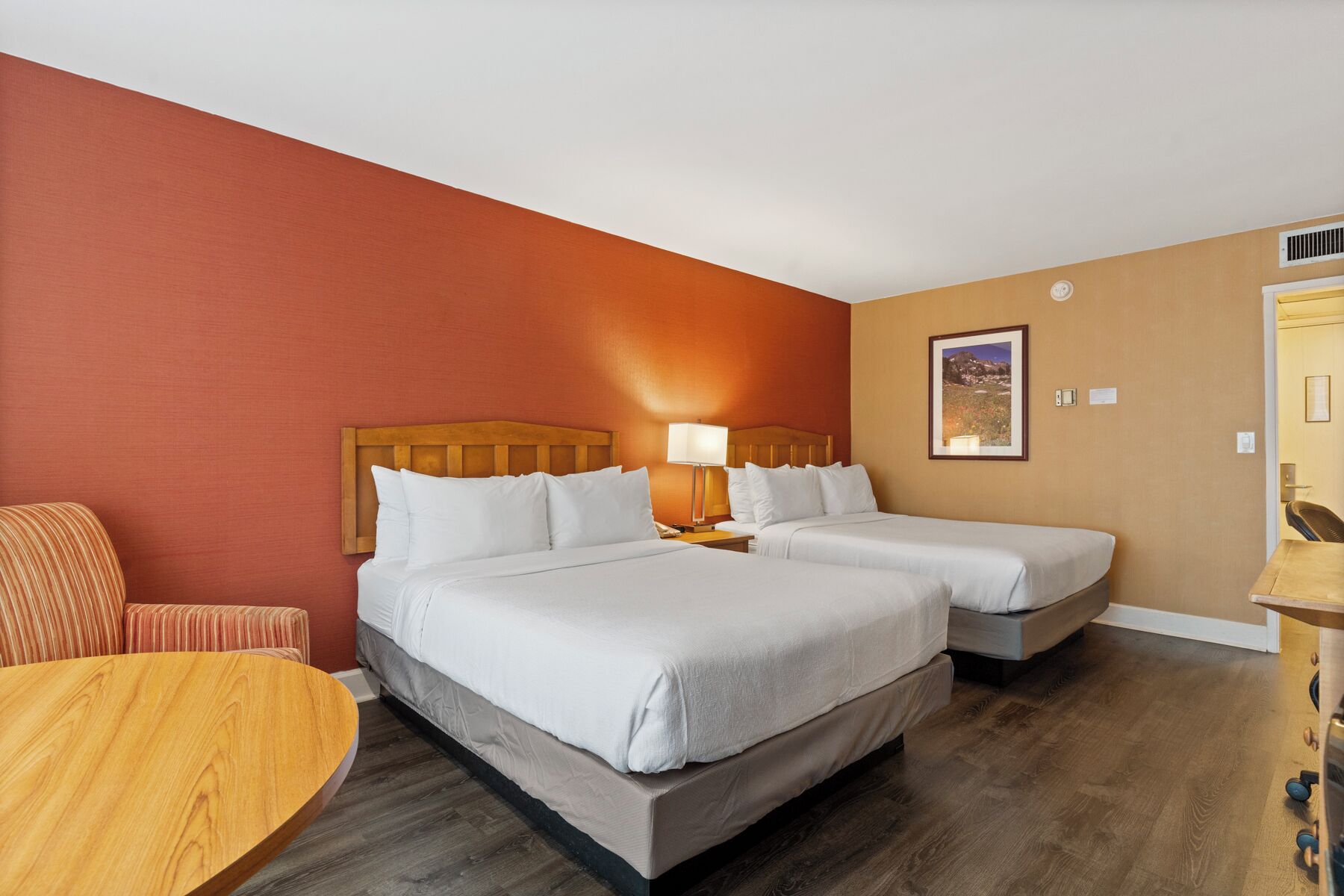 Habitación de hotel con dos camas, una silla de madera, una mesita y un cuadro en la pared.