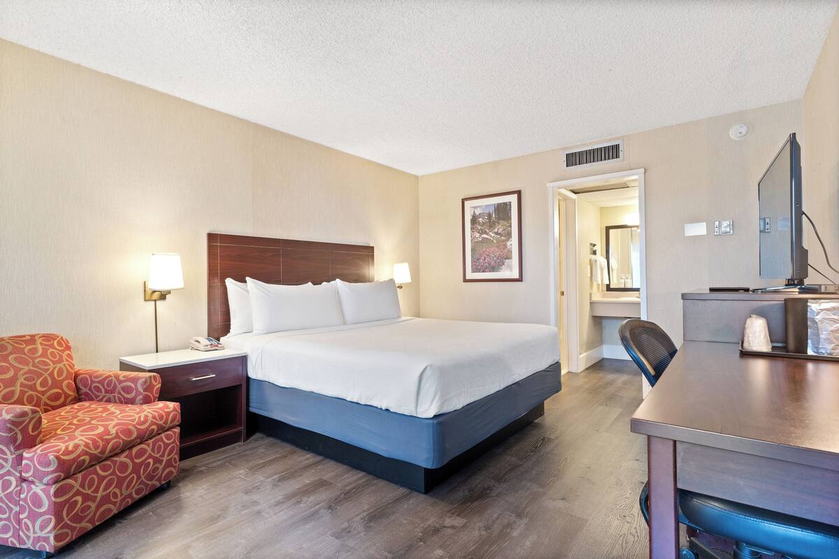 A hotel room with a bed, bedside table, lamp, chair, desk, and a television.