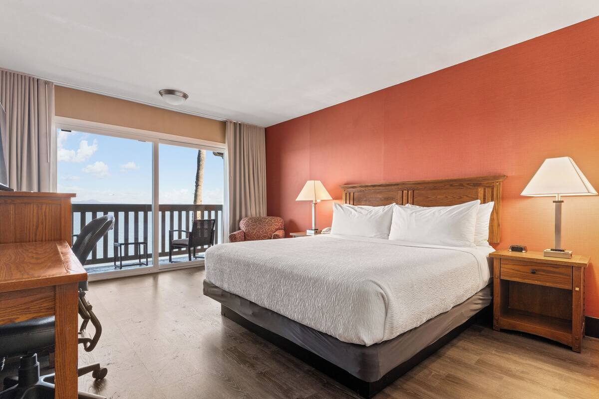 A hotel room with a large bed, two bedside lamps, a wooden nightstand, a desk with a chair, and a balcony with a view.