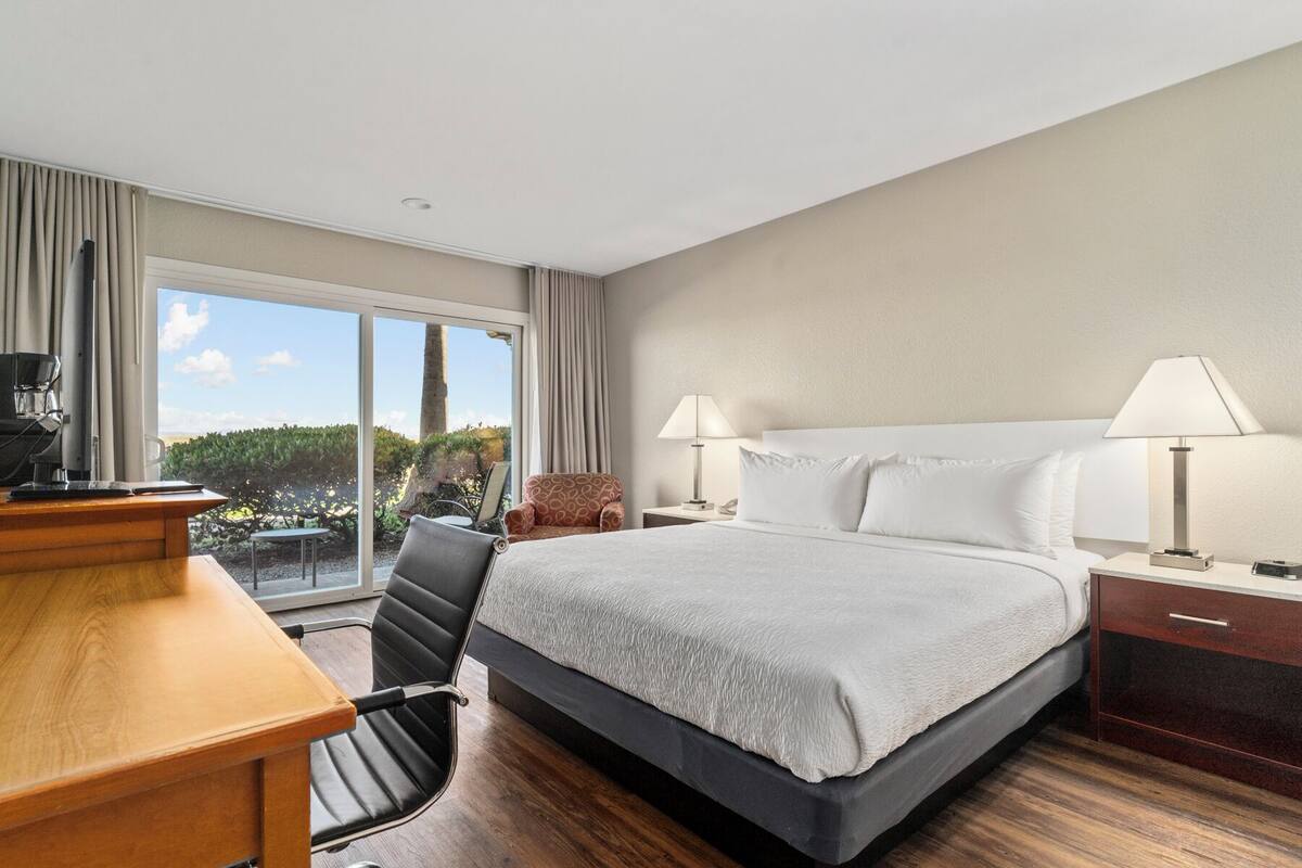 A hotel room with a large bed, a desk with a coffee machine, two bedside lamps, a chair, and a view of the outdoors.