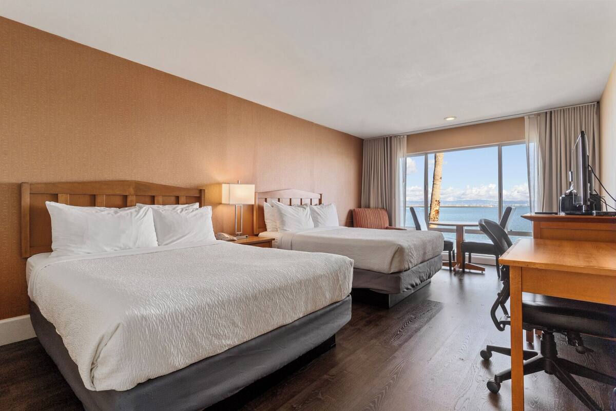 A hotel room with two beds, a desk, and a view of the sea.