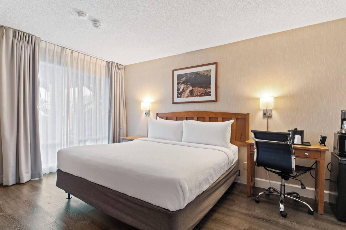 A hotel room with a large bed, a desk with a chair, a coffee machine, and a framed picture on the wall.