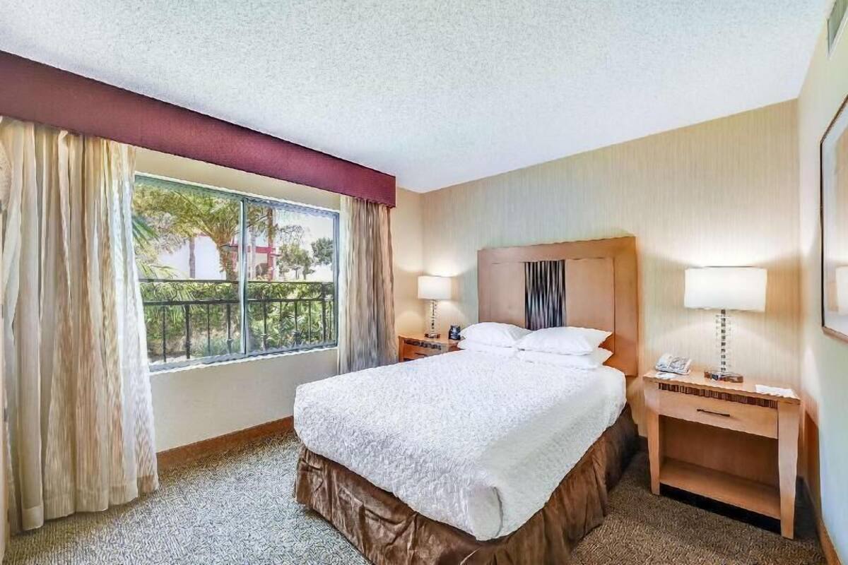 Habitación de hotel con una cama, dos mesitas de noche, una ventana con cortinas y vistas a zonas verdes.