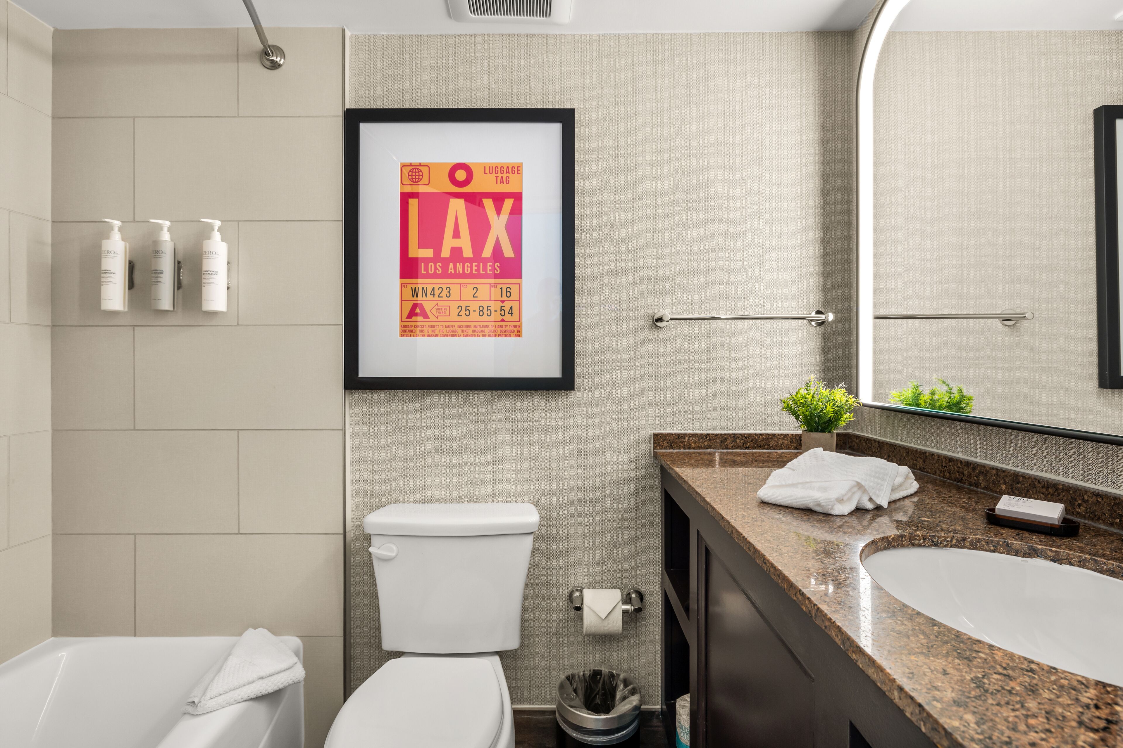A modern bathroom with a toilet, sink, and shower. There is a framed sign on the wall, a towel rack, and a small plant on the counter.
