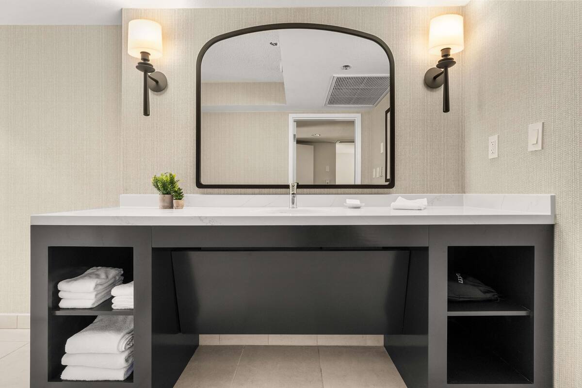 Un baño moderno con un espejo grande, dos luces empotradas, una encimera blanca y estantes con toallas dobladas.