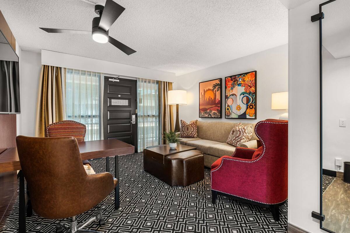 Habitación de hotel con comedor, sofá, sillón rojo, ventilador de techo y obras de arte en la pared.