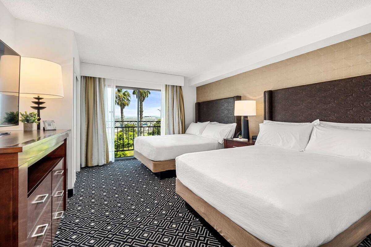 Habitación de hotel con dos camas, un escritorio y un balcón con vista a palmeras.