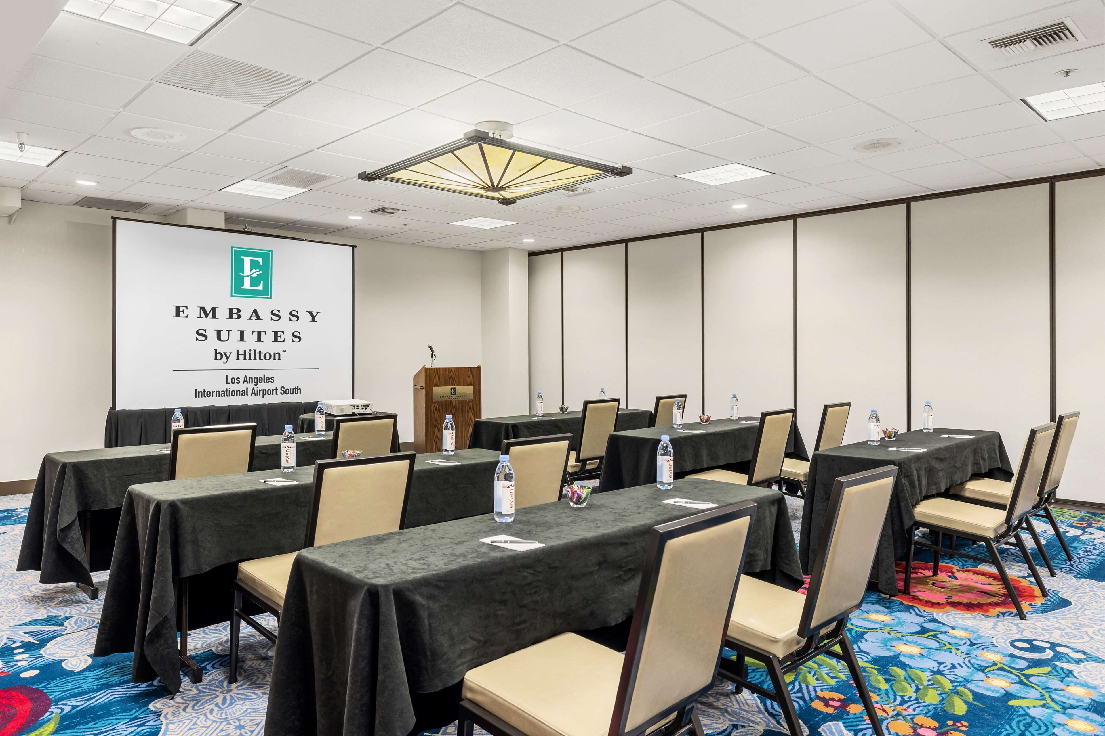 Una sala de conferències amb taules, cadires i una pantalla de projecció que mostra 'Embassy Suites by Hilton'.