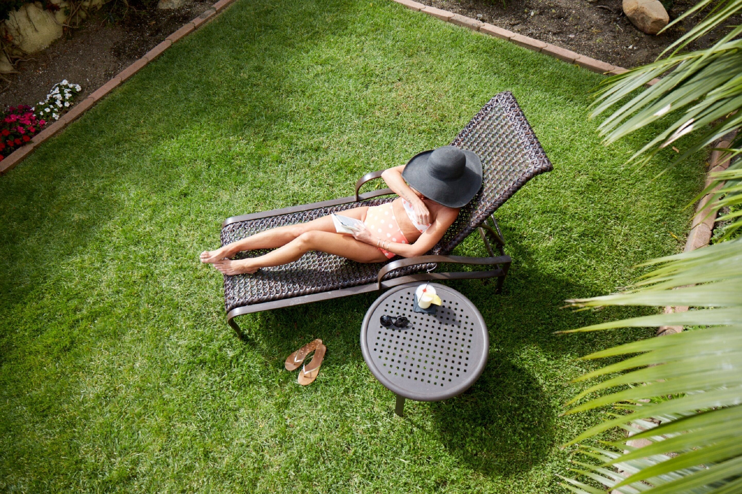 A person lying on a lounge chair in a garden area with a small table and a pair of sandals beside them.
