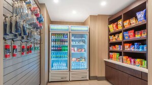 Un área de venta automática con dos dispensadores de bebidas, un expositor mural de artículos de cuidado personal y estantes repletos de aperitivos y bebidas.