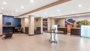Un moderno vestíbulo de hotel con mostrador de recepción, dos estaciones de trabajo con ordenador, un área de descanso y un jarrón decorativo sobre una mesa.