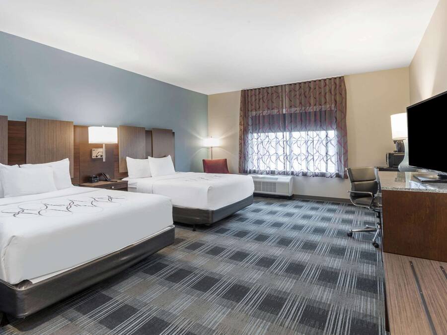 A hotel room with two beds, a desk, a television, and a large window with curtains.