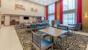 Un vestíbulo de hotel con varias mesas y sillas para comer, una zona de recepción con mostrador y televisor, y amplias ventanas con cortinas rojas.