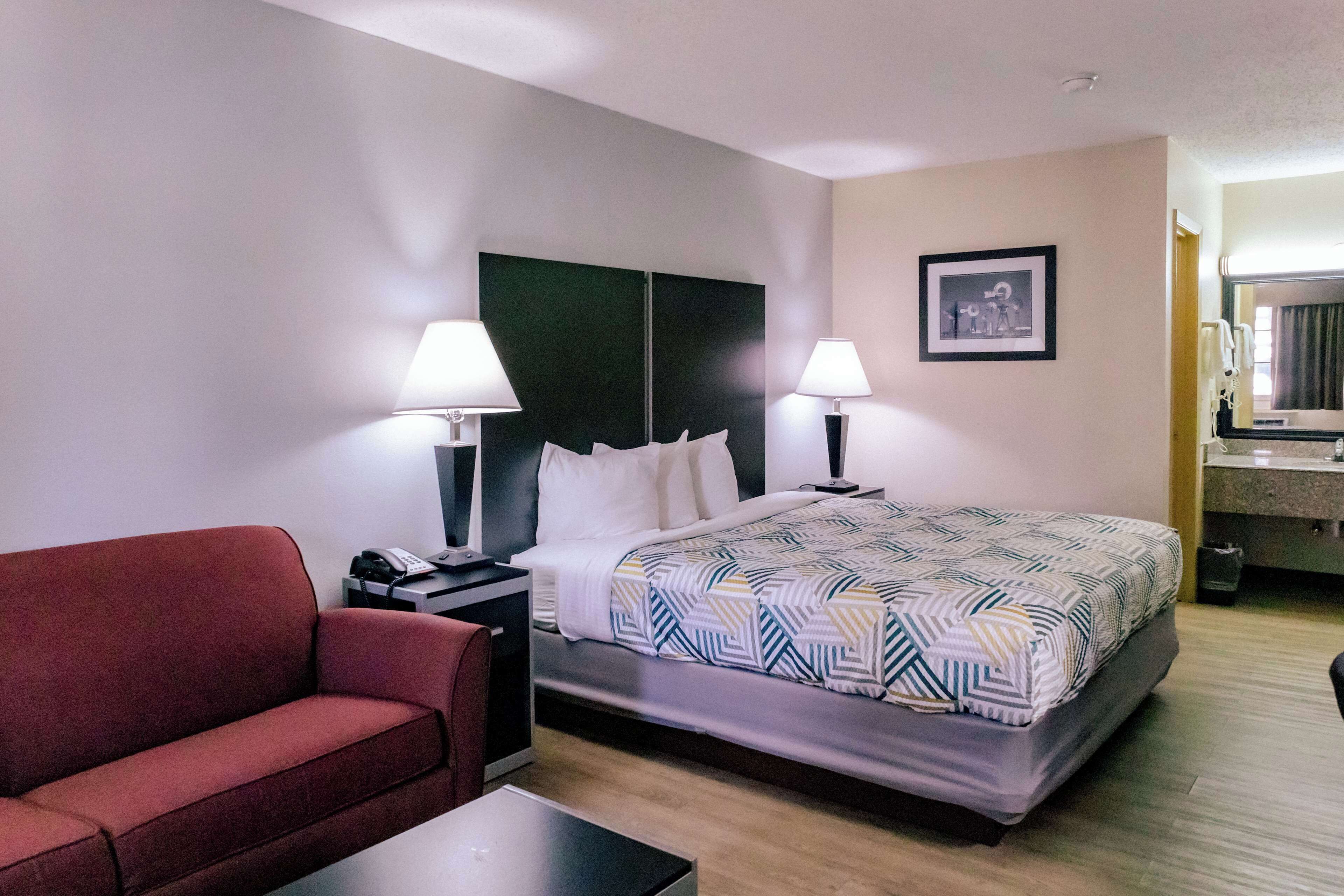 Quarto de hotel com cama, duas luminárias laterais, um sofá vermelho, uma mesa pequena e um quadro pendurado na parede.