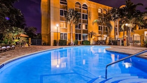 Una piscina con superficie de agua azul cristalina, rodeada por una terraza pavimentada y un edificio con grandes ventanales y palmeras.