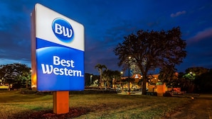 Letrero de Best Western con logotipo azul y blanco, rodeado de césped y árboles, con un edificio al fondo.
