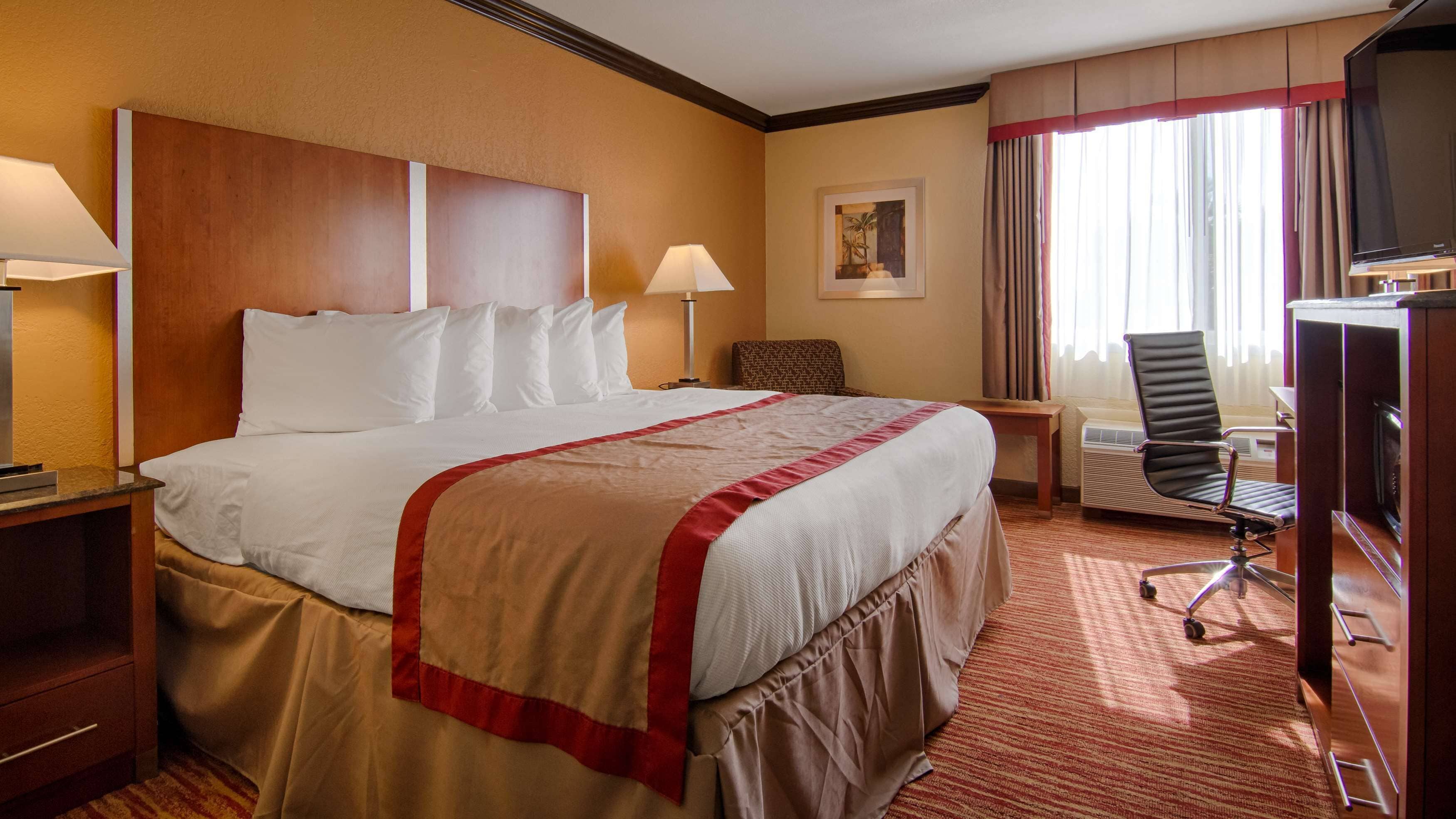 Ein Hotelzimmer mit einem großen Bett, einem Schreibtisch mit Lampe, einem Sessel, einem Fernseher und einer Klimaanlage.