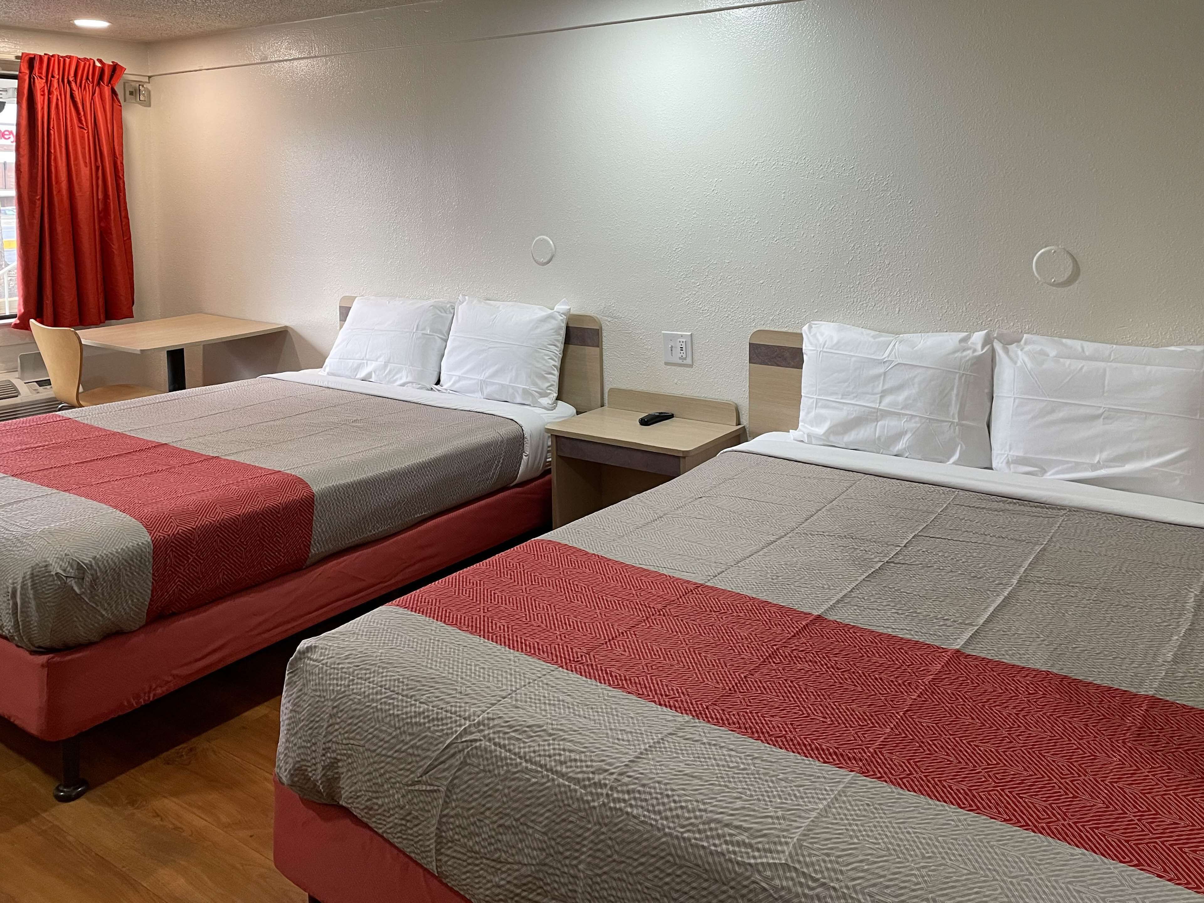 A hotel room with two beds, each with a red and grey bedspread, white pillows, and a wooden nightstand between them.