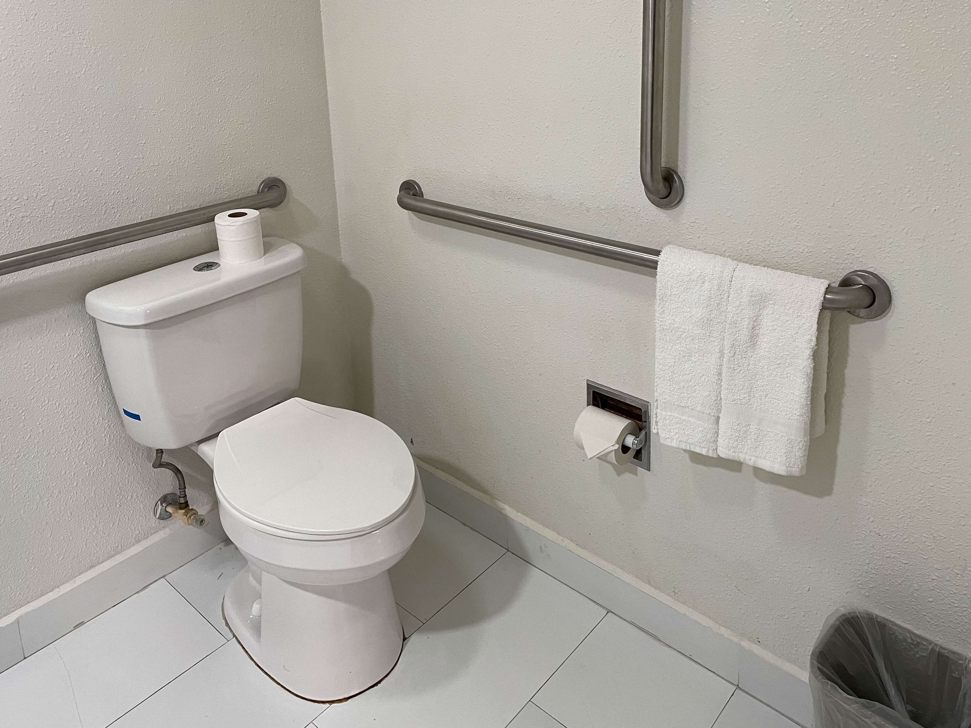 A standard bathroom with a toilet, a paper towel dispenser, a towel rack, and a trash bin.