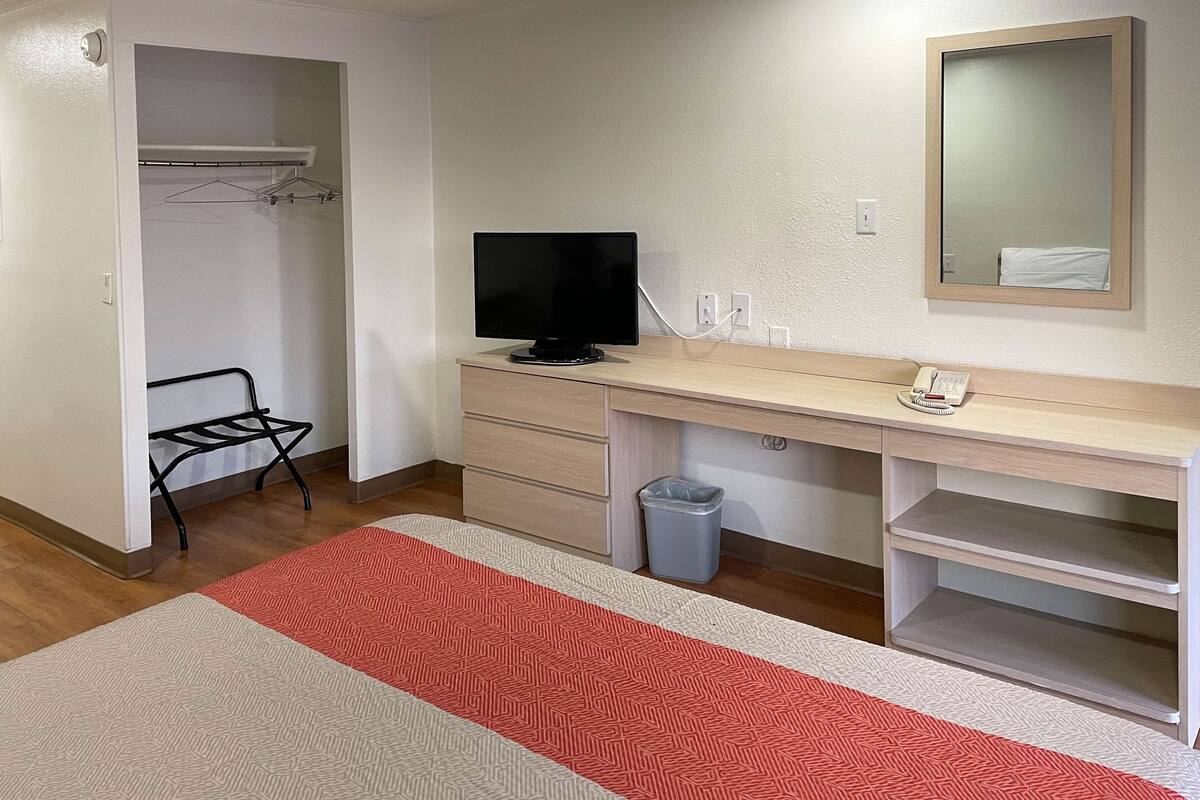 Ein Hotelzimmer mit einem Holztisch, einem Fernseher, einem Telefon, einem Papierkorb und einem Kleiderschrank mit Kleiderstange.