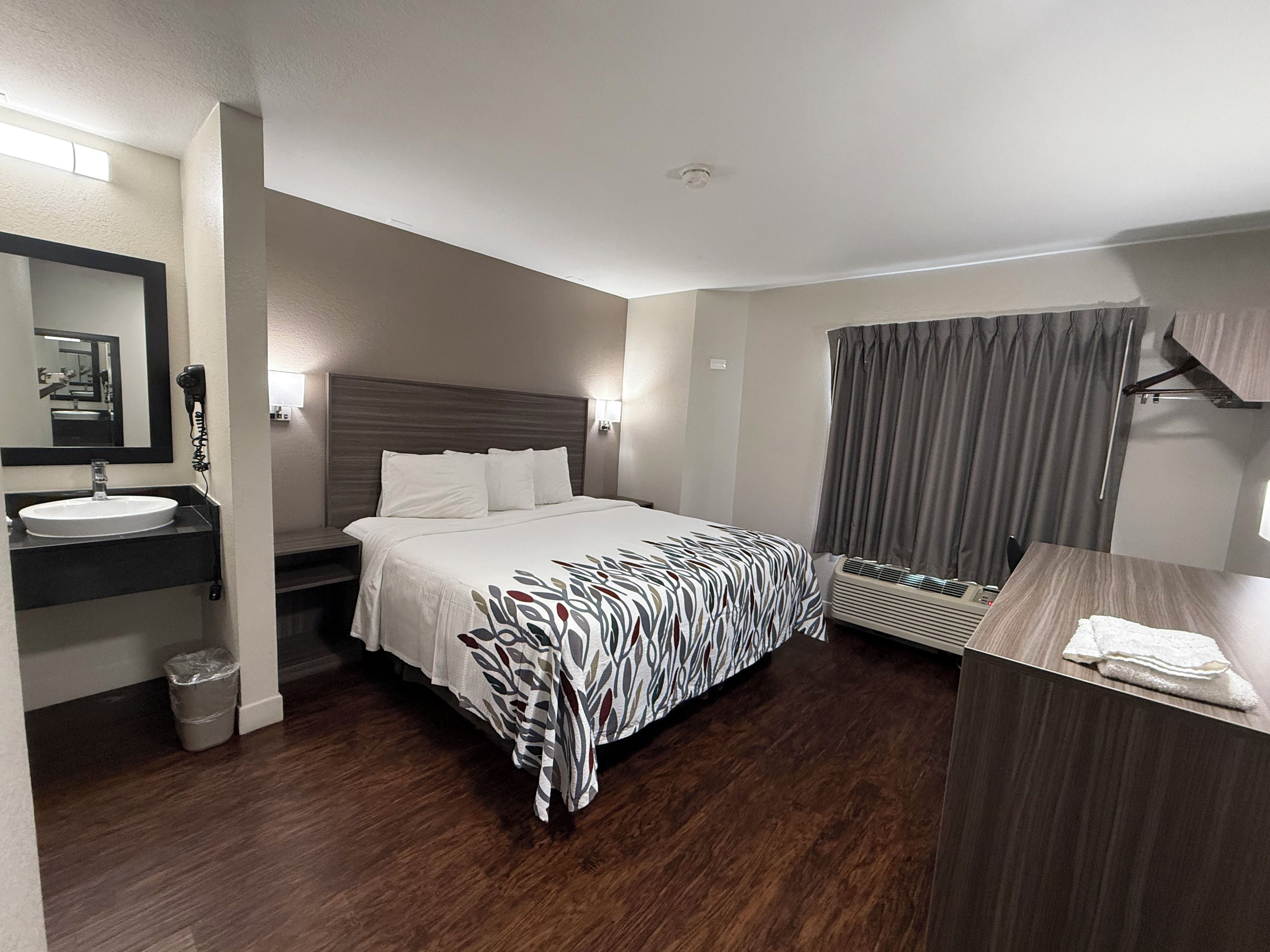 A hotel room with a bed, a bedside table, a mirror, a sink, and a window with curtains.