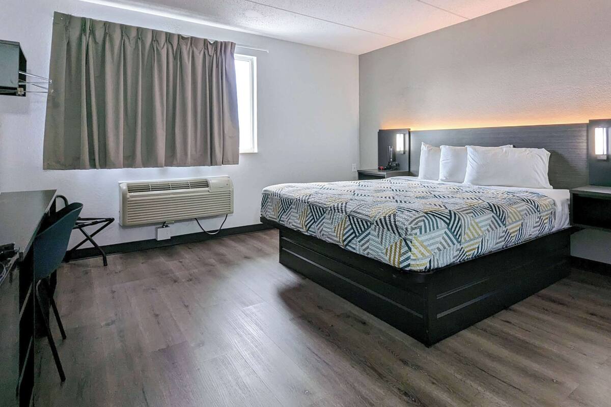 A hotel room with a bed, bedside tables, a desk, an air conditioner, and a window with curtains.