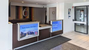 A reception desk with a large number '6' on the wall, two digital screens displaying a booking message, and a tiled floor. - Motel 6 Danvers, MA - Boston North (Danvers)