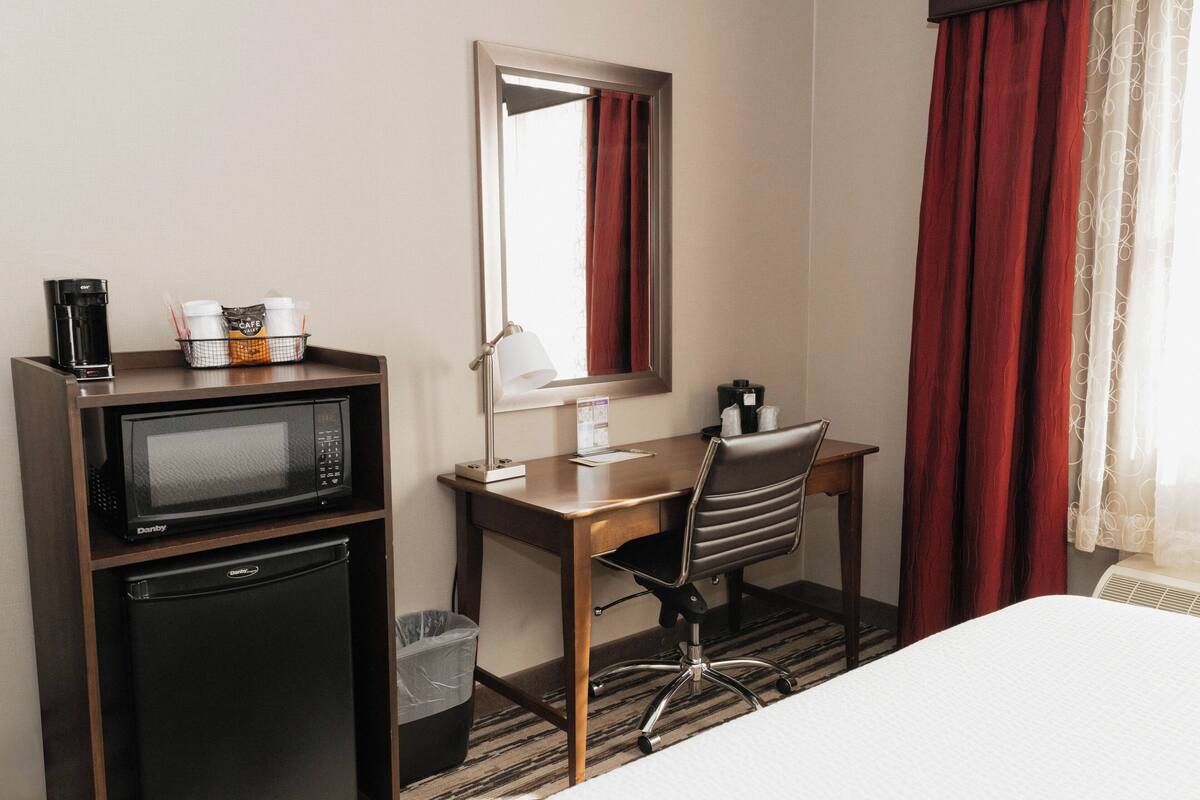 A hotel room with a bed, a desk with a chair, a microwave, a refrigerator, a trash bin, and a window with curtains.