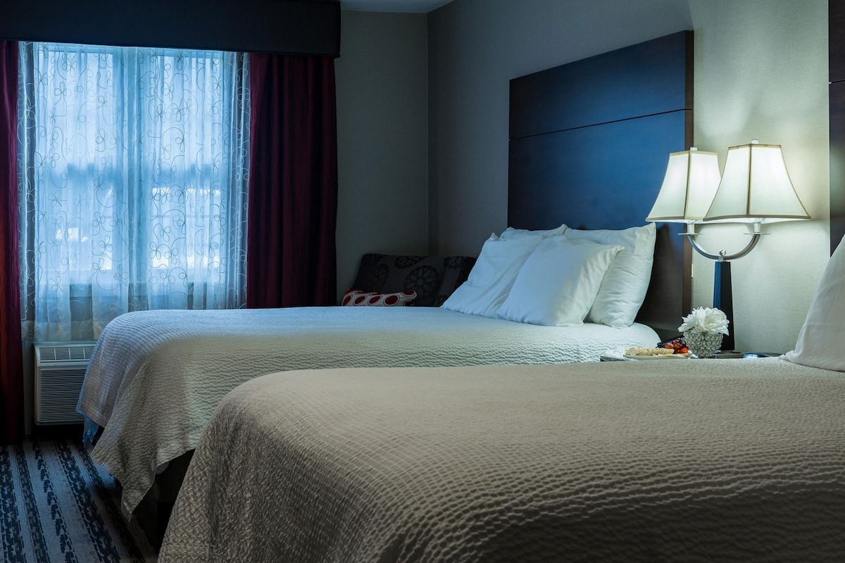 A hotel room with two beds, a large window with curtains, a bedside table with a lamp, and a wall-mounted headboard.