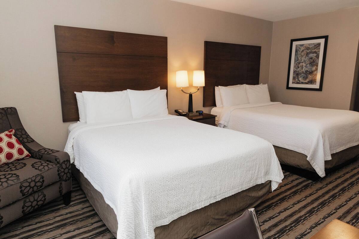 A hotel room with two beds, a nightstand with a lamp, a chair, and a framed picture on the wall.