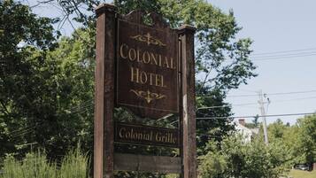 Un letrero de madera anuncia el Hotel Colonial, con un muro de piedra y flores rojas en primer plano.