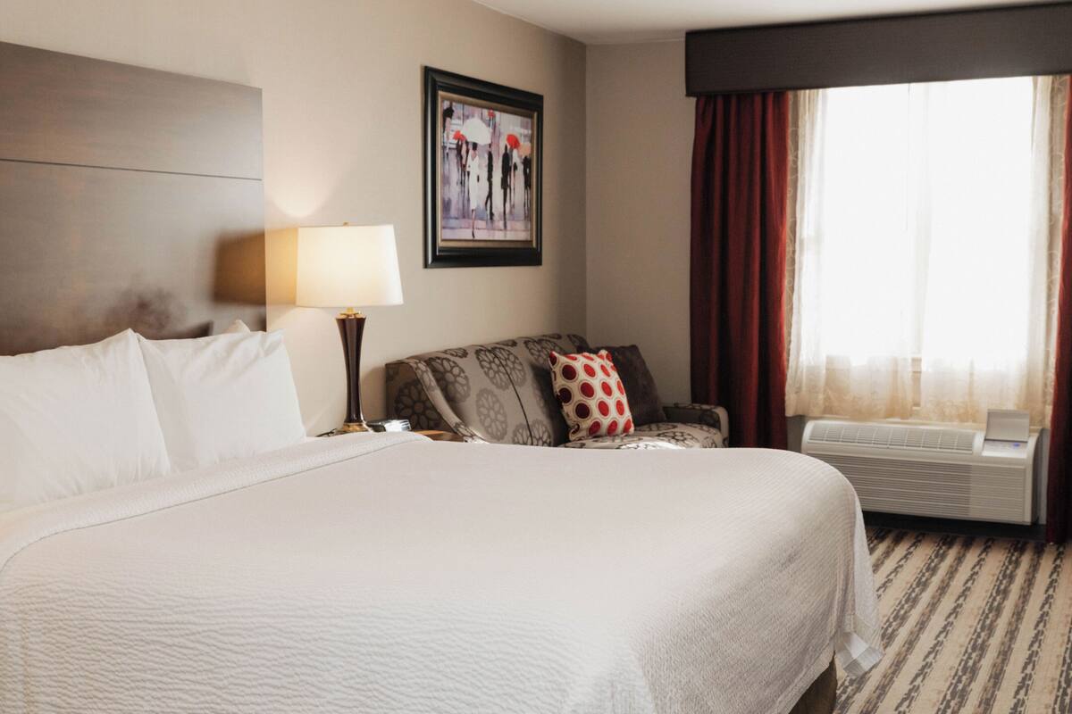 A hotel room with a large bed, a sofa, a desk with a lamp, a framed artwork on the wall, and a window with curtains.