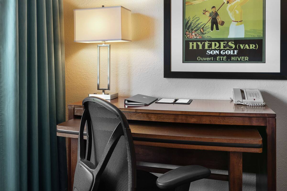 A hotel room with a wooden desk, a black office chair, a lamp, a framed poster, and a telephone.