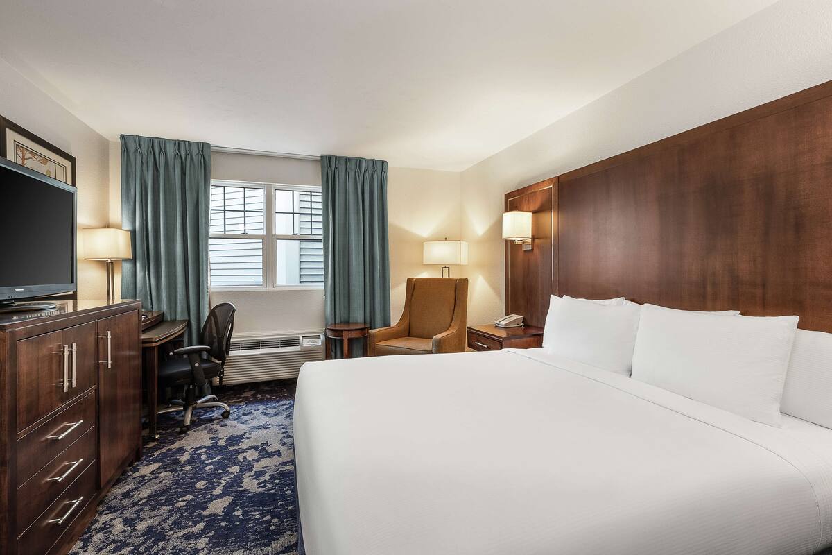 A hotel room with a bed, a desk with a chair, a television, a lamp, and a window with curtains.