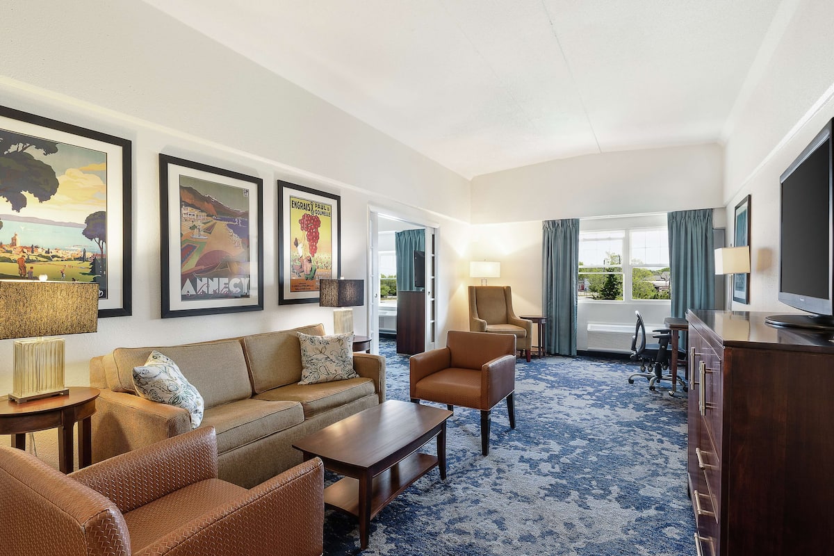 A hotel room with a sofa, armchairs, a coffee table, a TV, and framed artwork on the wall.