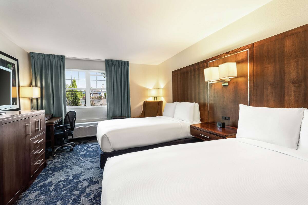 A hotel room with two beds, a desk, a chair, and a large window with curtains.