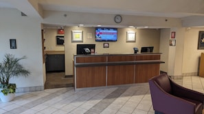 A reception area with a wooden counter, a digital screen displaying information, a clock, and a potted plant. - Red Roof Inn San Antonio - Airport (San Antonio)