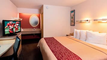 A hotel room with a bed, a desk with a television, a mirror, and a wall-mounted light fixture.