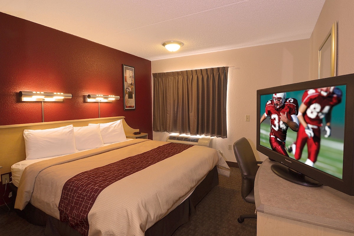 Quarto de hotel com cama, televisão, escrivaninha e janela com cortinas.