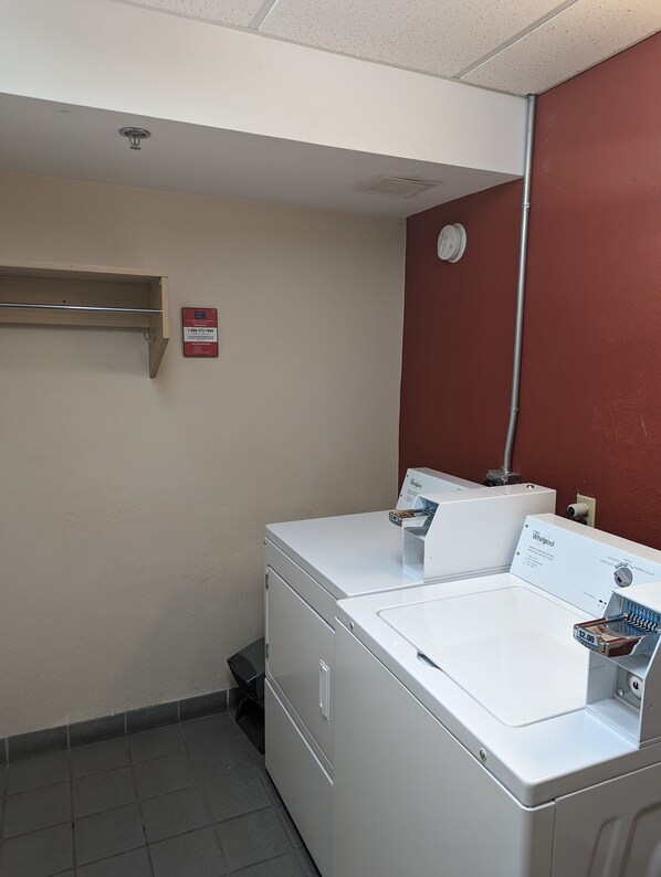 A laundry room with a front-loading washer and dryer, a metal shelf, a red wall, and a white ceiling. - Red Roof Inn San Antonio - Airport (San Antonio)