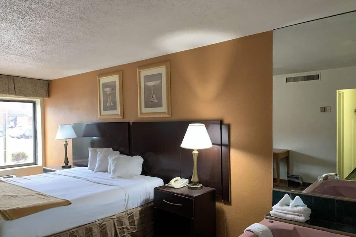 A hotel room with a bed, two bedside lamps, a nightstand, a telephone, a mirror, and a bathroom with a bathtub.