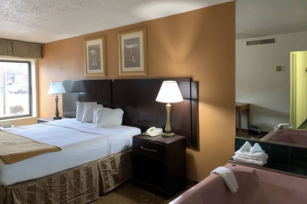 A hotel room with a large bed, two bedside lamps, a nightstand with a telephone, a mirror, and a bathroom with a sink and towel stack.