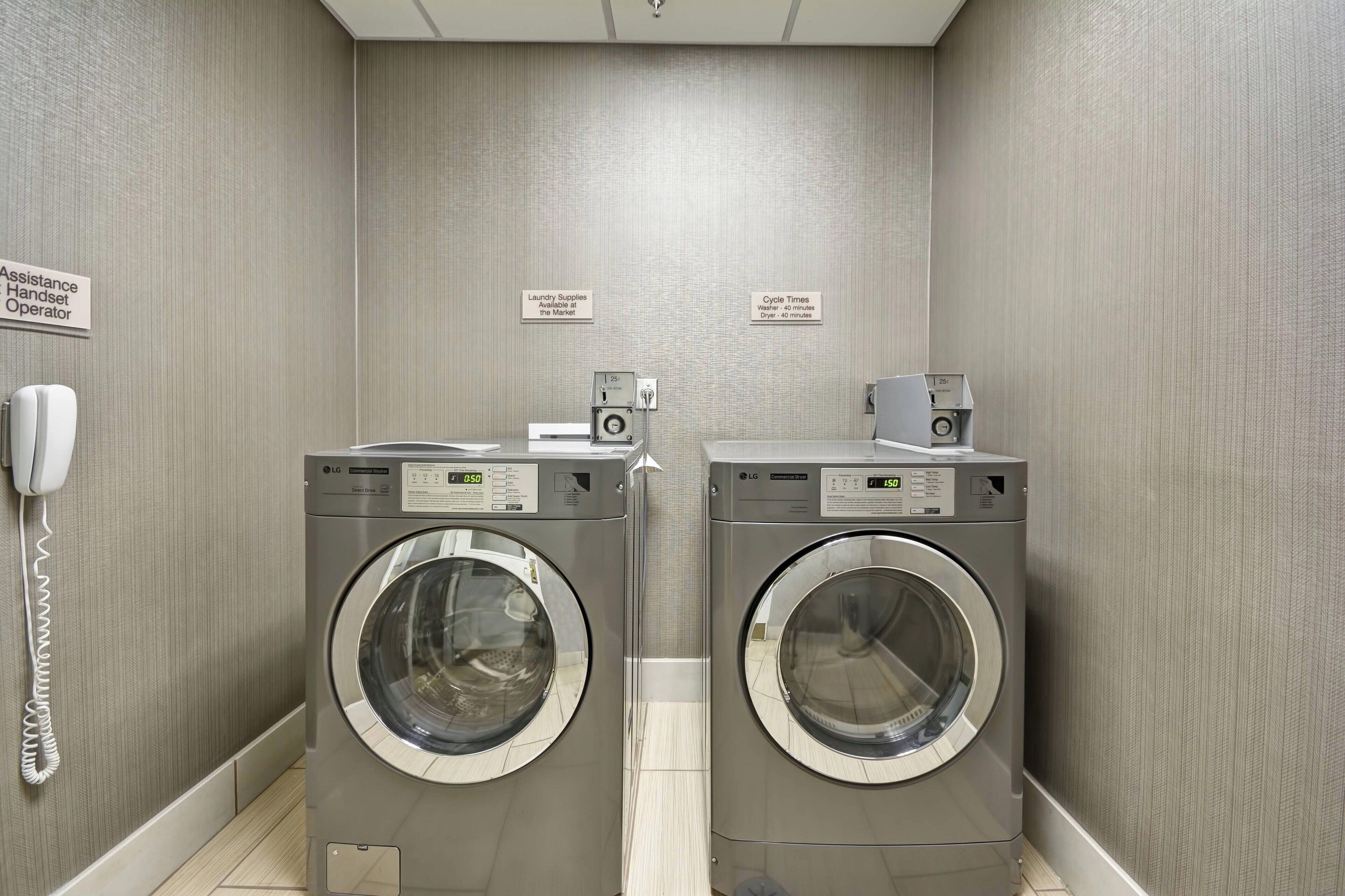 A laundry room with two front-loading LG washers, a phone, and signs indicating assistance and laundry supplies.