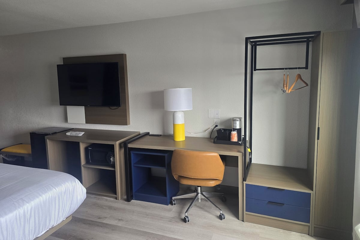 Habitación de hotel con cama, escritorio con silla, televisor, lámpara, cafetera y armario.