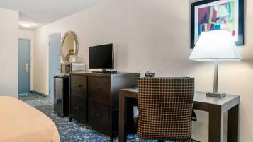 Una habitación de hotel con cama, escritorio con computadora, espejo, lámpara y un cuadro en la pared.