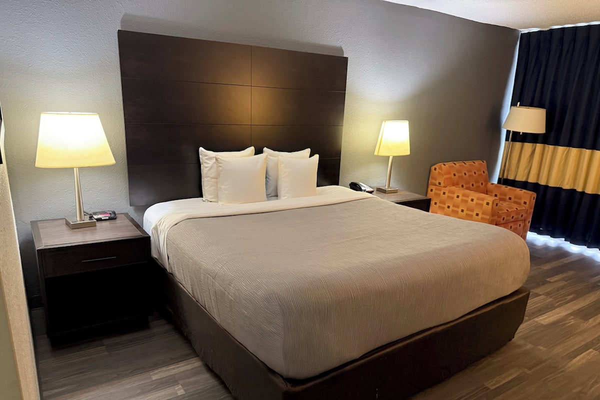 Une chambre d’hôtel avec un grand lit, deux tables de chevet avec des lampes, une tête de lit en bois, une couvre-lit à motifs, un plancher en bois et un fauteuil à motifs.
